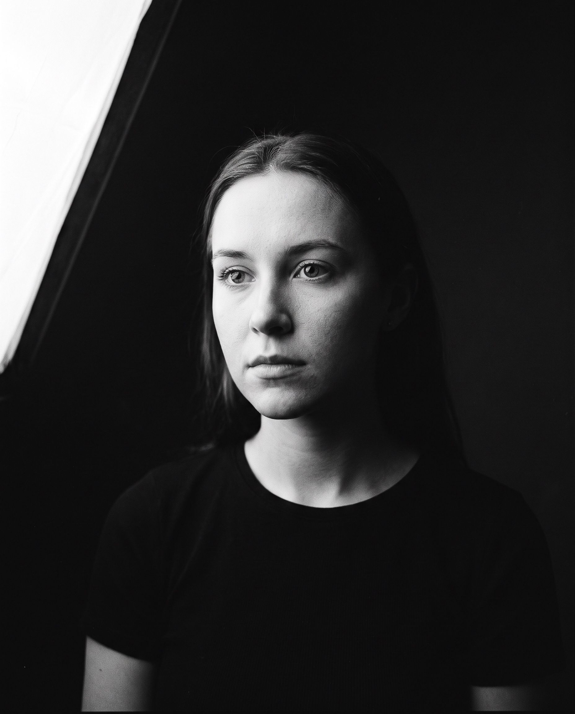 Black and white studio headshot of a young woman lit by a single softbox, deep shadow falling away behind her