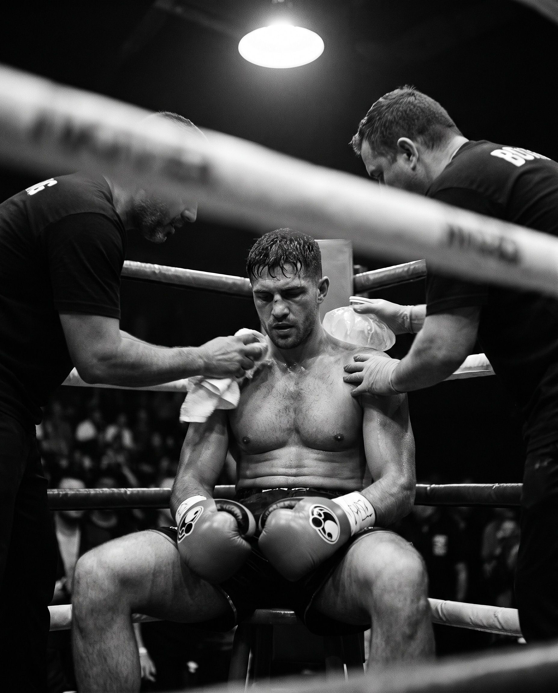 A boxer sitting in the corner between rounds under a hard overhead light, ropes crossing the foreground